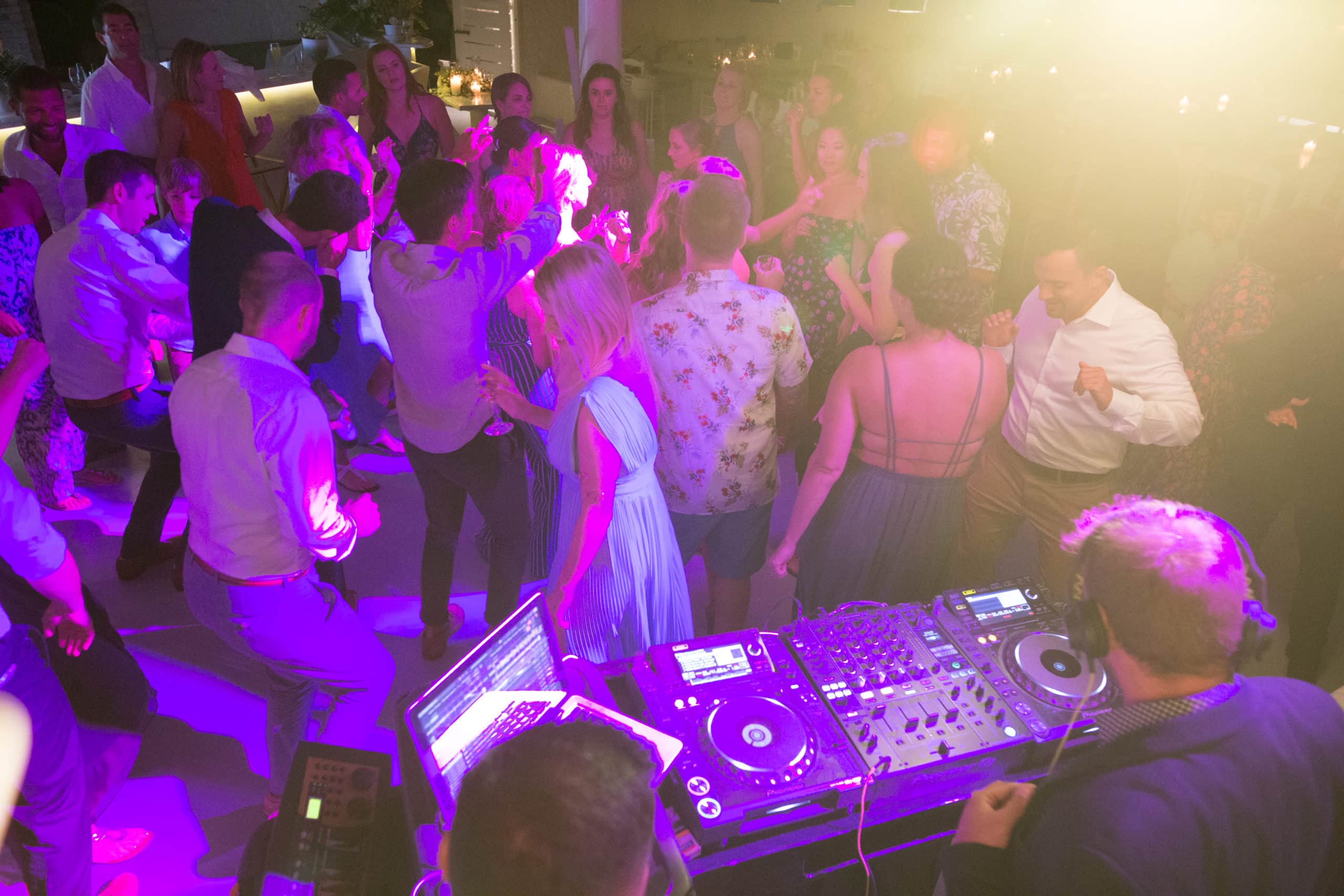 View from the DJ booth: Pioneer DJ decks in front of a dancing wedding crowd lit in magenta