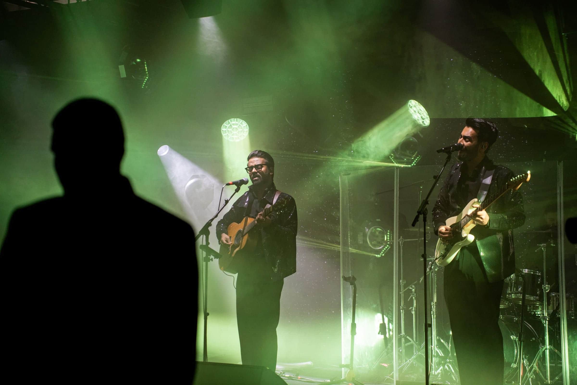 Two musicians performing on stage under green and white stage lighting, seen past a silhouetted guest