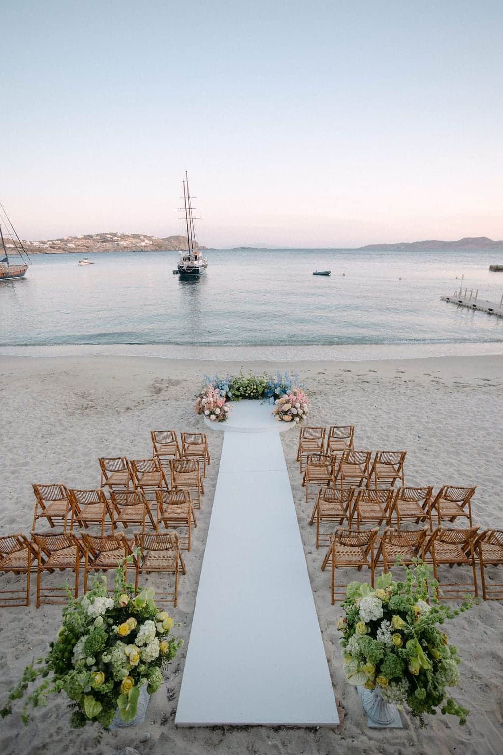 Beach wedding ceremony set with a white aisle and flowers, facing a bay with a sailing ship