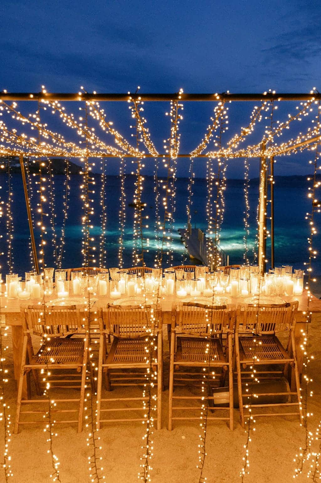 Long dinner table under a canopy of fairy lights at dusk by the sea