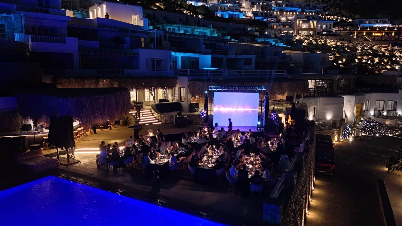 Night-time dinner on a hillside hotel terrace with an LED screen on a truss beside the pool