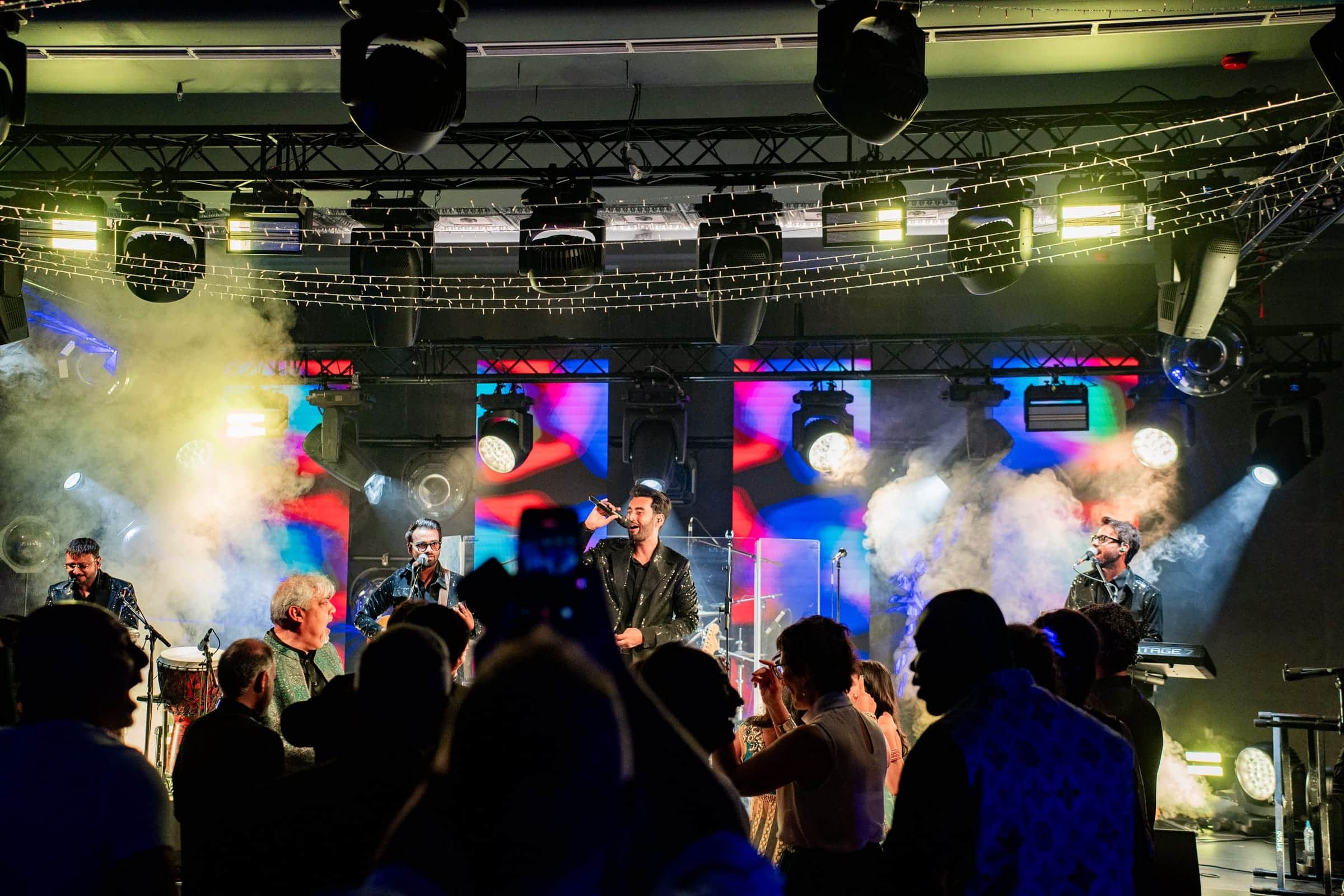 Live band on a stage with a lighting truss, moving heads, LED panels and haze as guests dance in front
