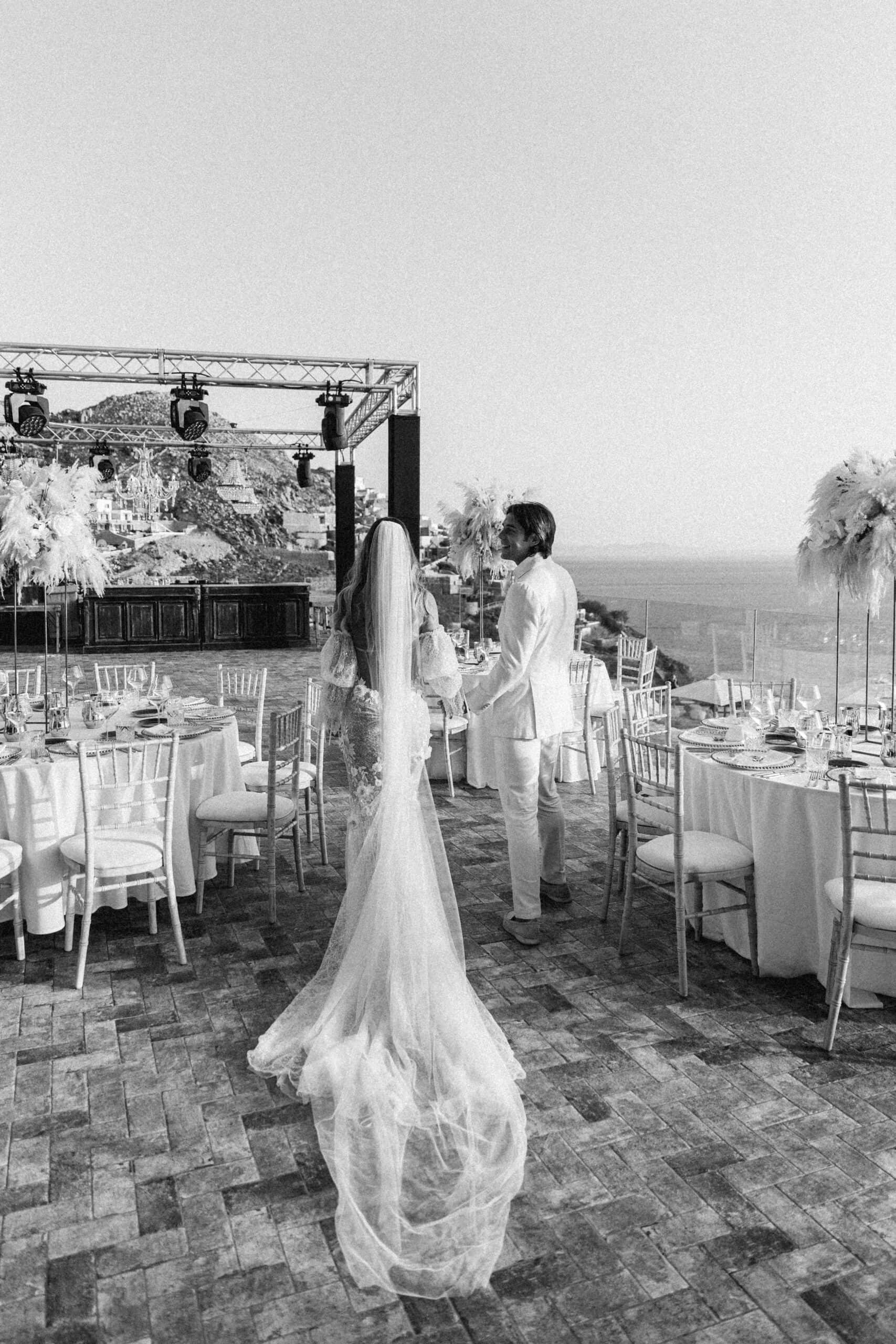 Black-and-white photo of the bride walking past reception tables beneath a lighting truss in Mykonos