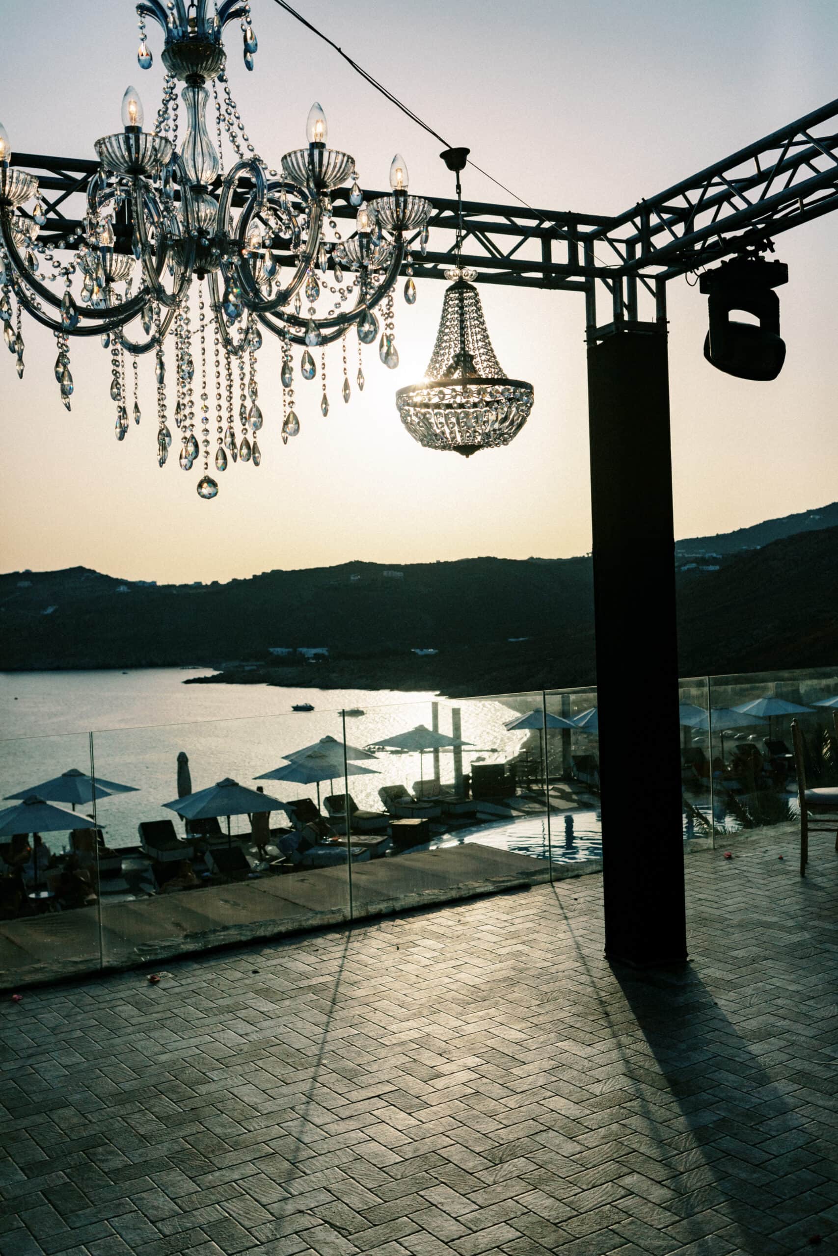 Crystal chandeliers and a moving-head light hung from a lighting truss above a Mykonos wedding terrace at sunset