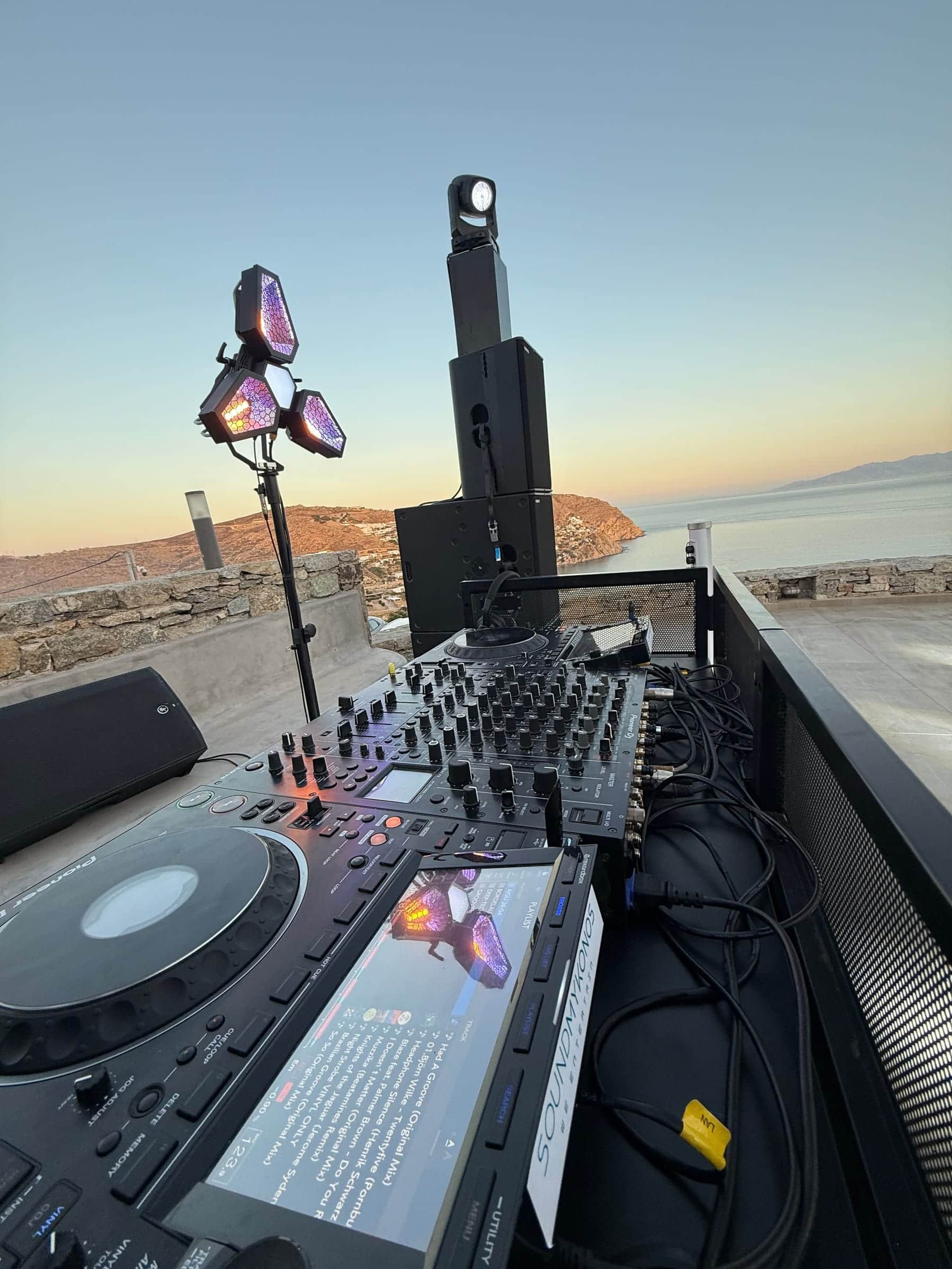 SoundMykonos DJ booth at sunset above the sea in Mykonos: Pioneer CDJ-3000, DJ mixer, a Portman Mantis light and a speaker stack topped by a moving head