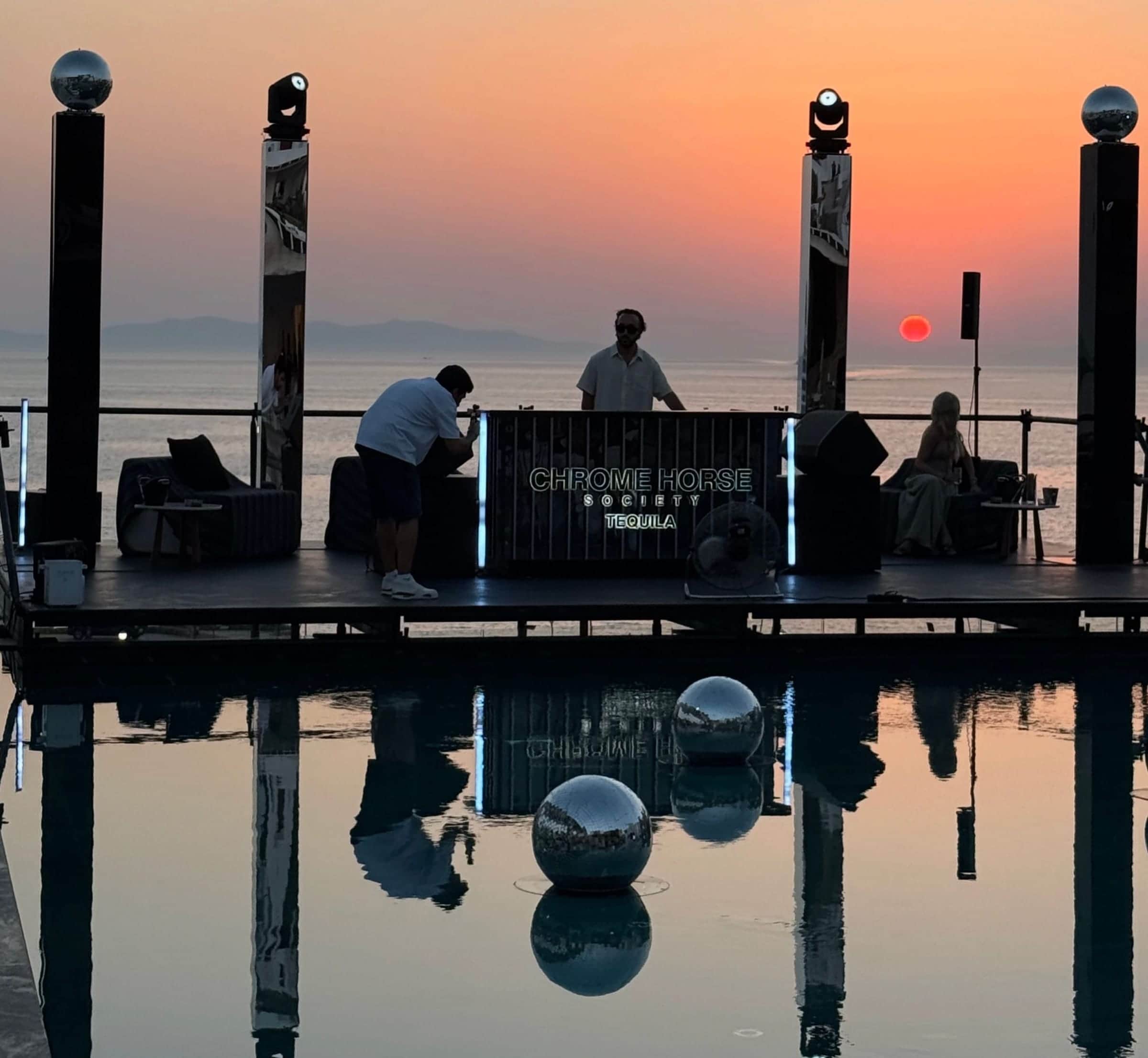DJ booth on a stage above an infinity pool at sunset in Mykonos, framed by moving-head lights on posts