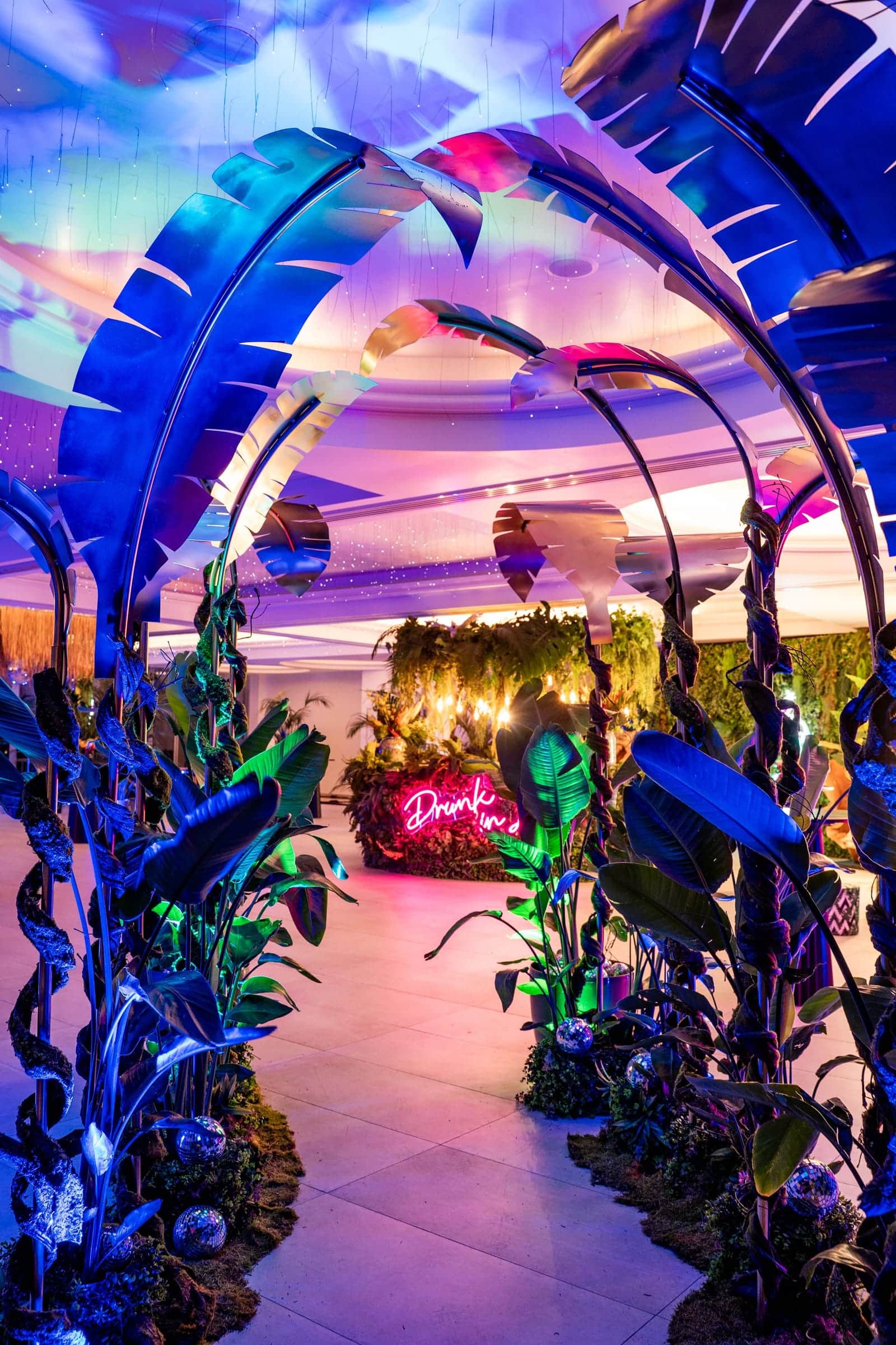 Tropical-themed event entrance with leaf arches and a neon sign under purple wash lighting